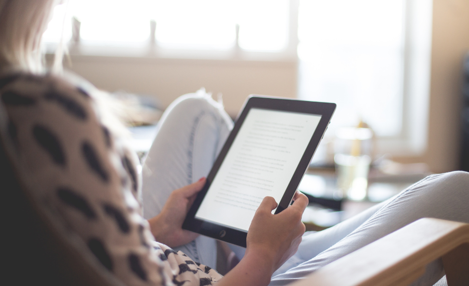 Personne assise dans un salon lumineux, lisant un texte sur une tablette électronique, avec un livre posé à proximité.