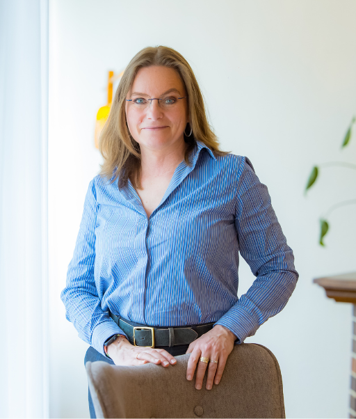 Delphine Malapert souriante aux cheveux mi-longs blonds, portant une chemise bleue à rayures et des lunettes, debout derrière une chaise dans un intérieur lumineux.