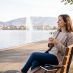 Une femme assise sereinement sur un banc au bord du lac Léman à Genève, tenant une boisson chaude et regardant au loin le Jet d'eau, illustrant l'apaisement, la régulation émotionnelle et la prise de recul.
